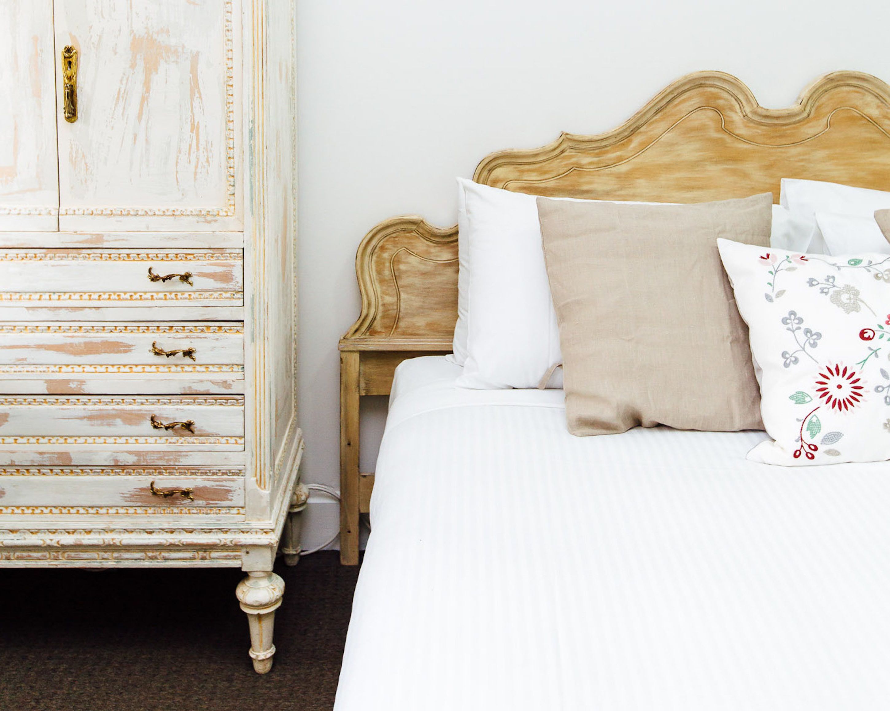 A bedroom with a white bed, a wooden nightstand, and a white dresser with gold handles.