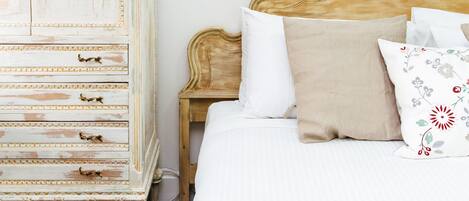 A bedroom with a white bed, a wooden nightstand, and a white dresser with gold handles.