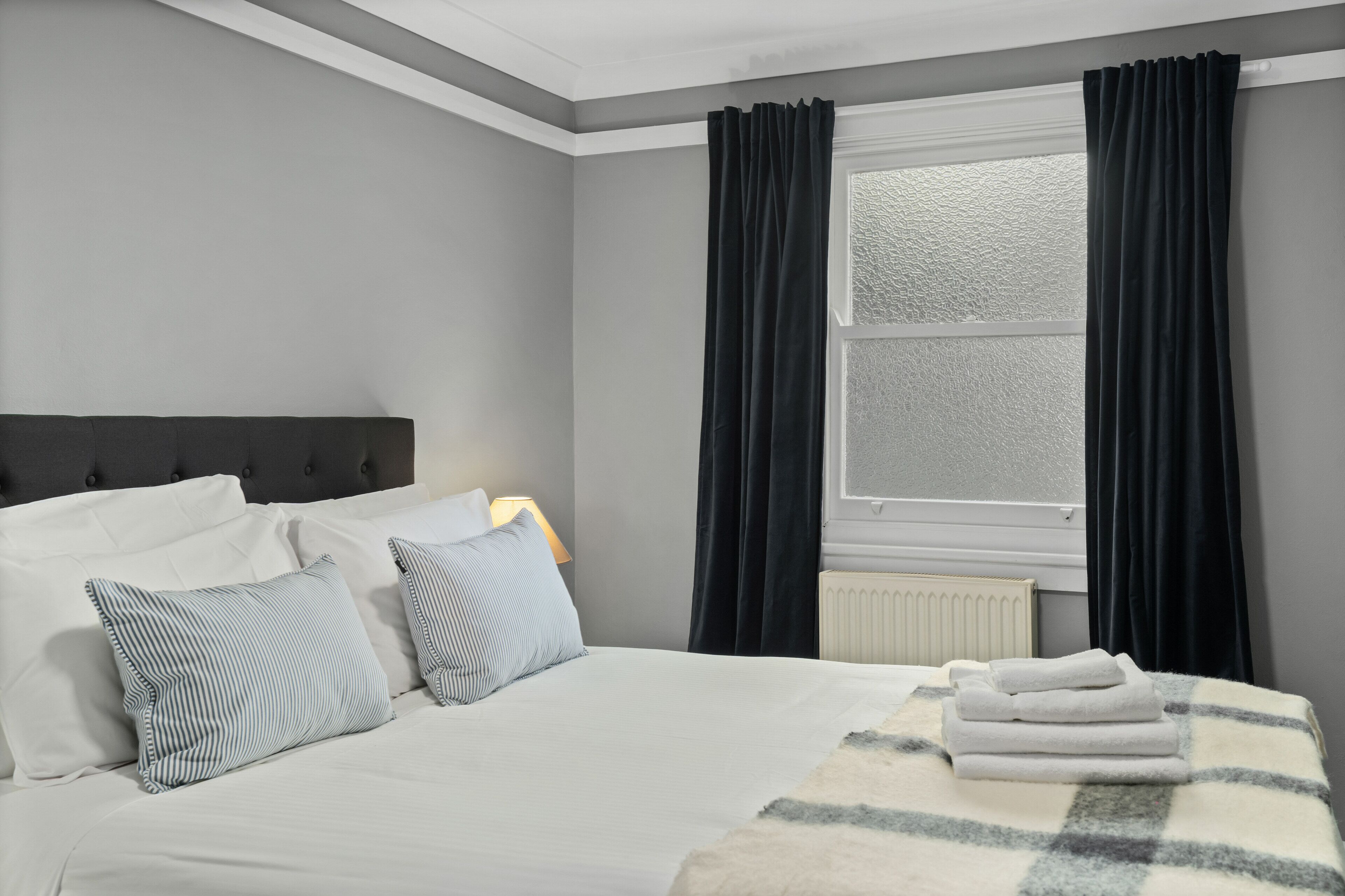 A neatly made bed with white linens and striped pillows, a white radiator, and dark curtains over a window.