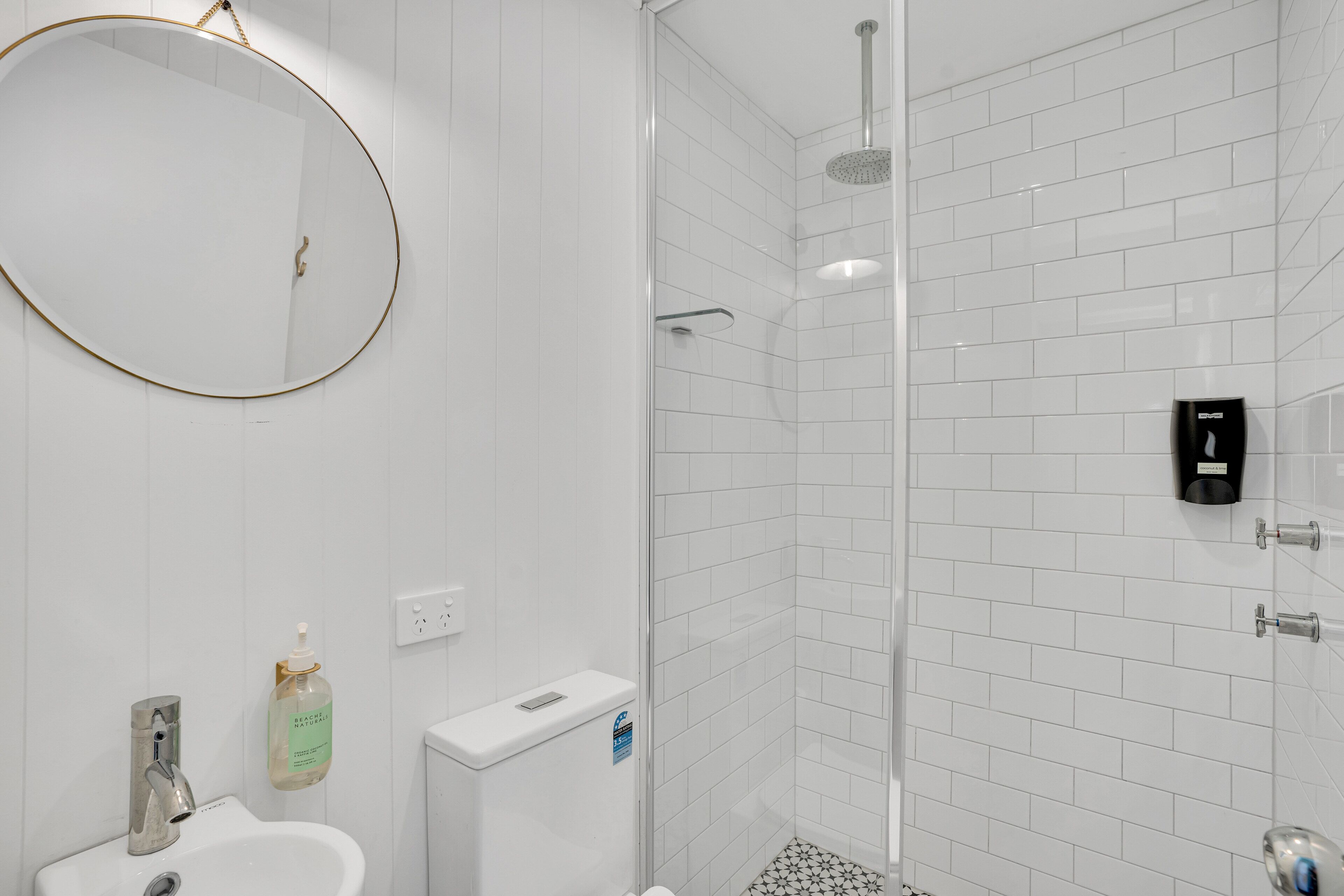 A bathroom with a round mirror, a sink, a toilet, and a shower area with white tiled walls and floor.
