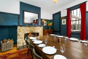 A dining room with a long wooden table set for a meal, a stone fireplace, and a large window with red curtains. - Bundanoon Hotel (Bundanoon)