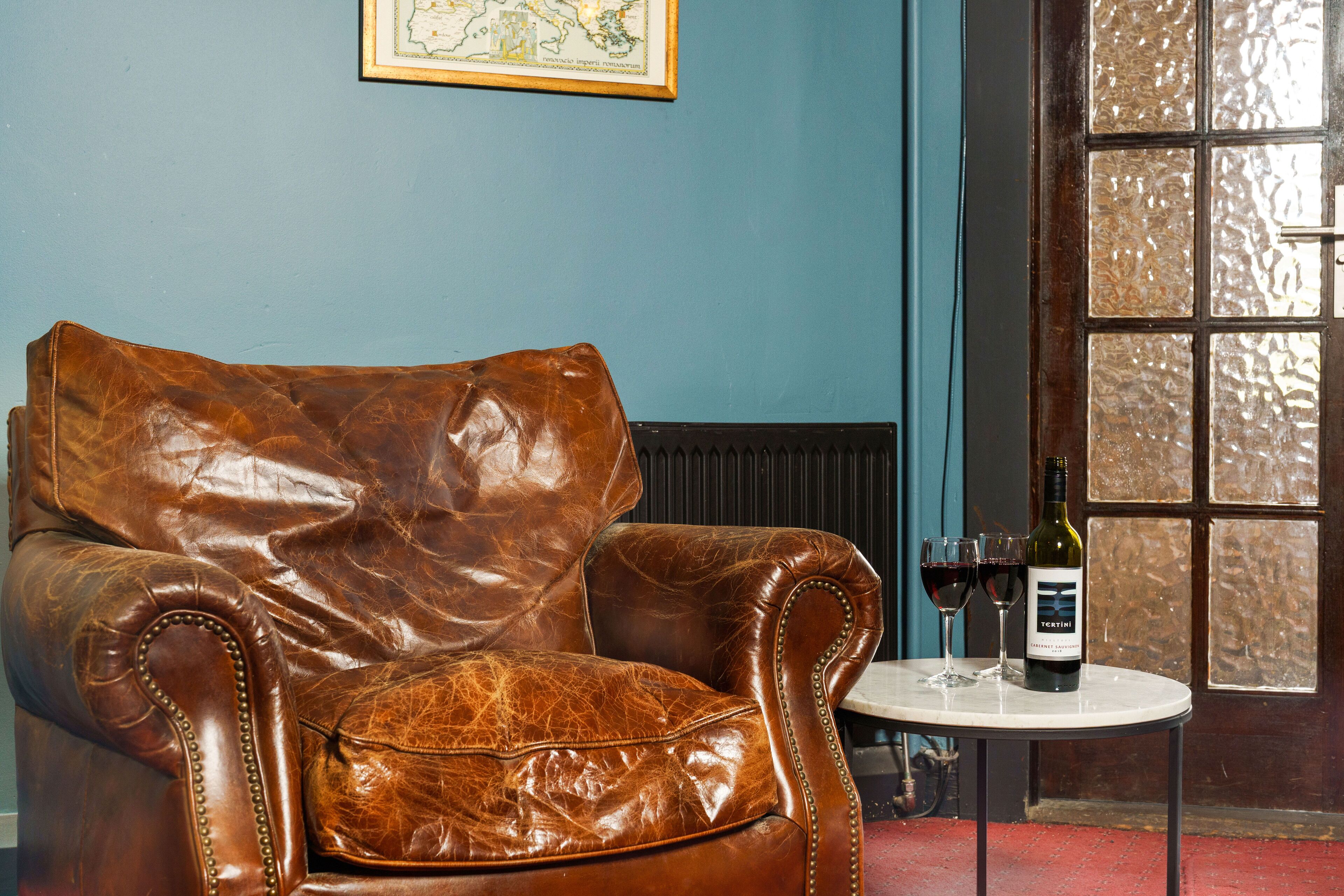 Un sillón de cuero marrón con un respaldo alto, una mesita redonda con una botella de vino y dos copas, y un mapa enmarcado en la pared.