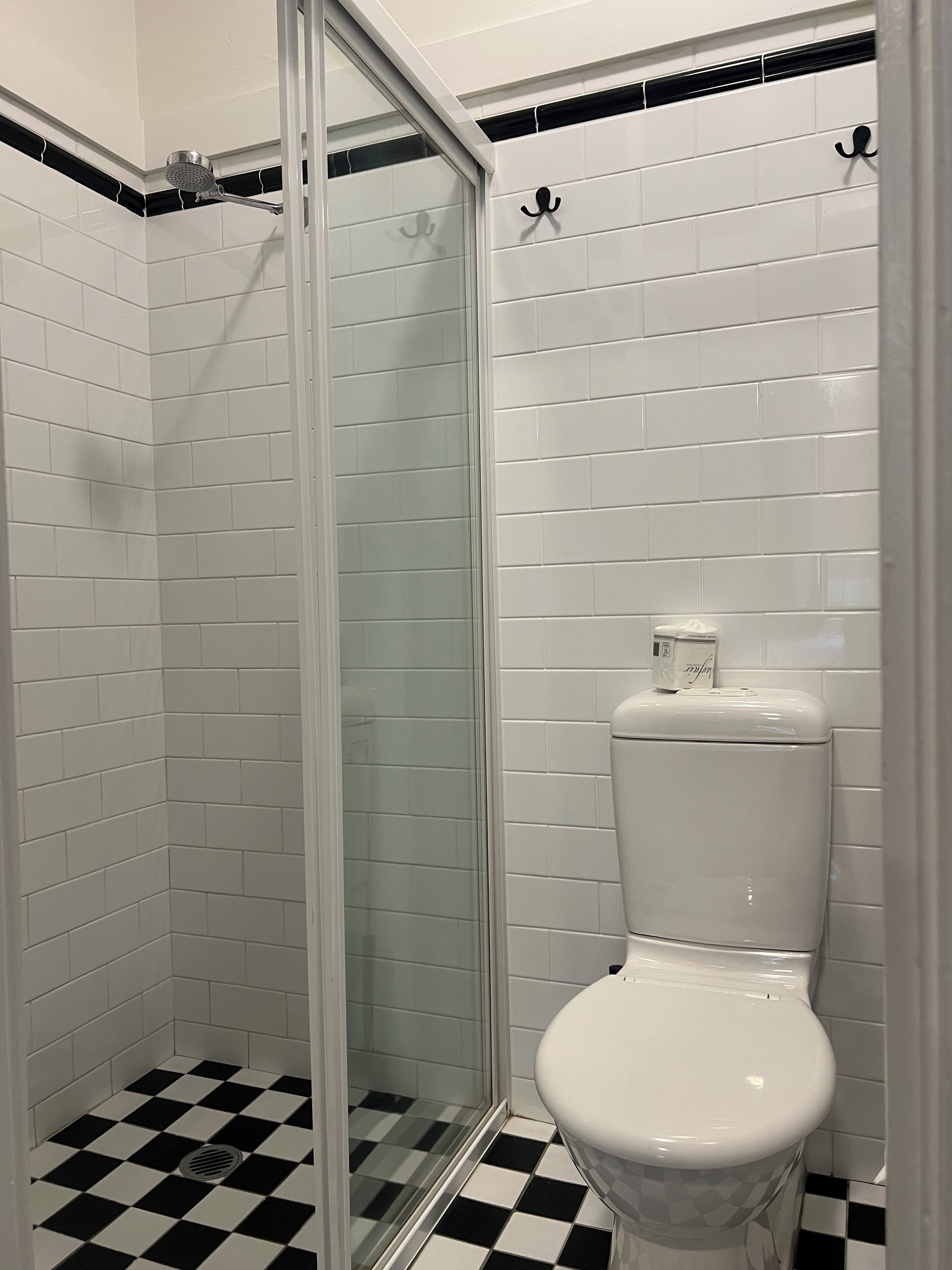 A bathroom with white subway tiles, a black and white checkered floor, a glass shower enclosure, and a toilet with a paper towel holder.