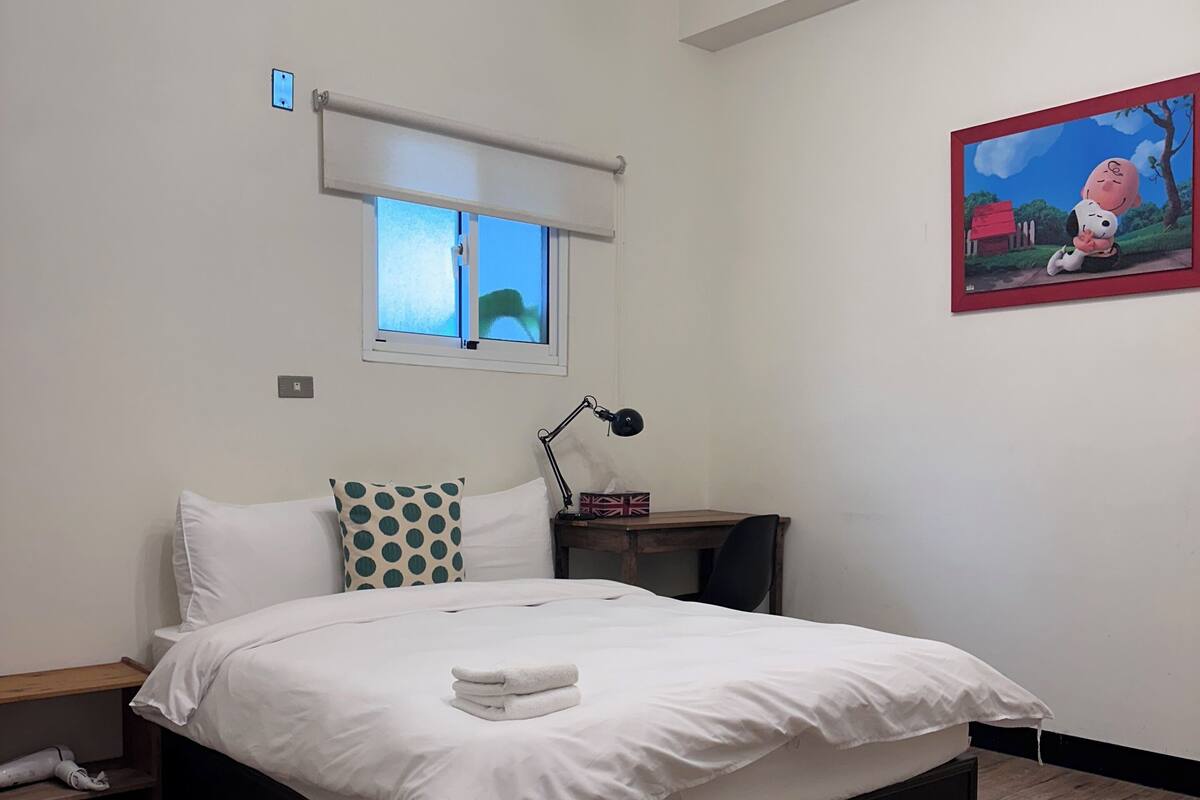 A neatly made bed with white linens and a folded towel, a bedside table with a lamp, a window with blinds, and a framed picture on the wall.