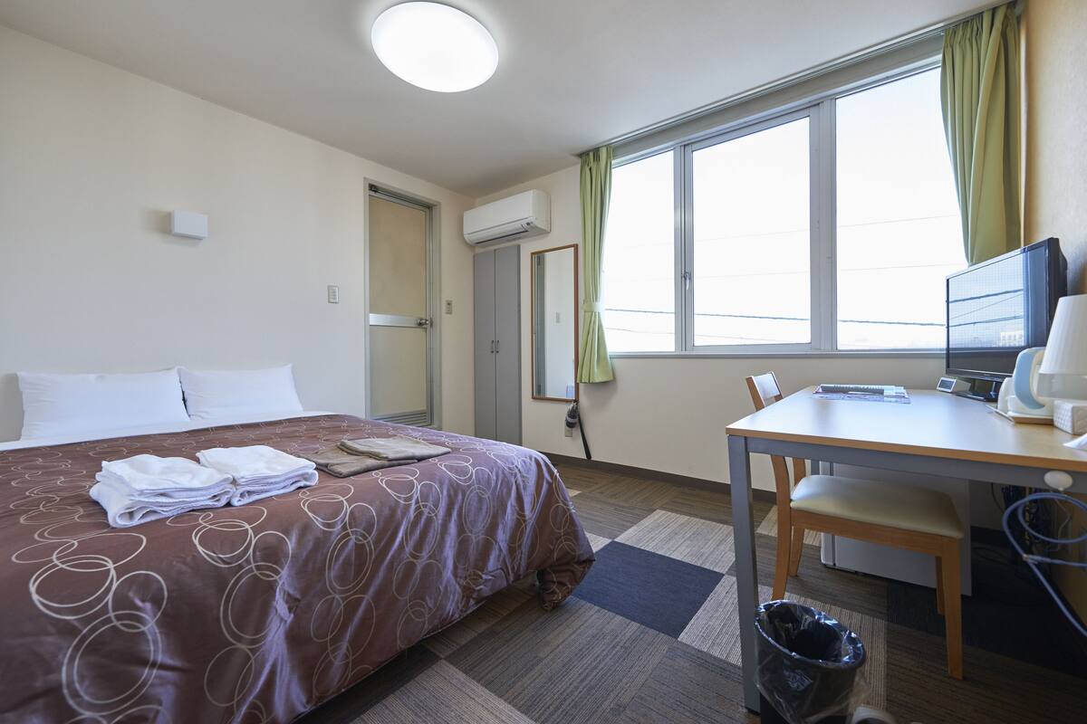 A hotel room with a bed, a desk with a chair, a computer, and a window with curtains.