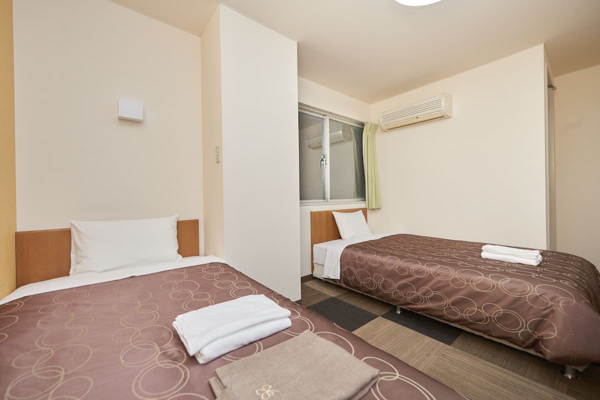 A hotel room with two beds, each with a brown patterned bedspread and white pillows. There is a window with a green curtain, an air conditioning unit on the wall, and a small white object on the wall.