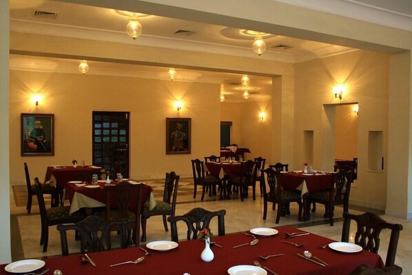 A dining area with multiple tables and chairs, arranged in a spacious hall with wall-mounted lighting. - King's Abode (Kumbhalgarh)