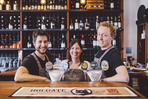 Un bar con tres personas detrás de la barra, una variedad de botellas en estantes y un cartel que dice "HOLGATE".