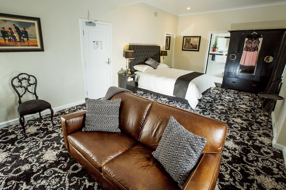 A hotel room with a brown leather sofa, a large bed with a dark headboard, a wardrobe, and a patterned carpet.