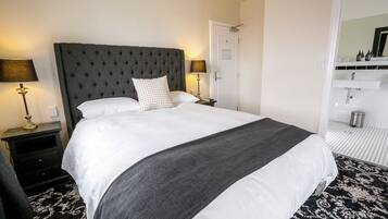 Een net opgemaakt bed met een grijs hoofdbord, wit beddengoed en een grijze plaid. Er staat een nachtkastje met een lamp en een klein tafeltje naast het bed. Achtergrond toont een badkamer met een witte wastafel en spiegel.