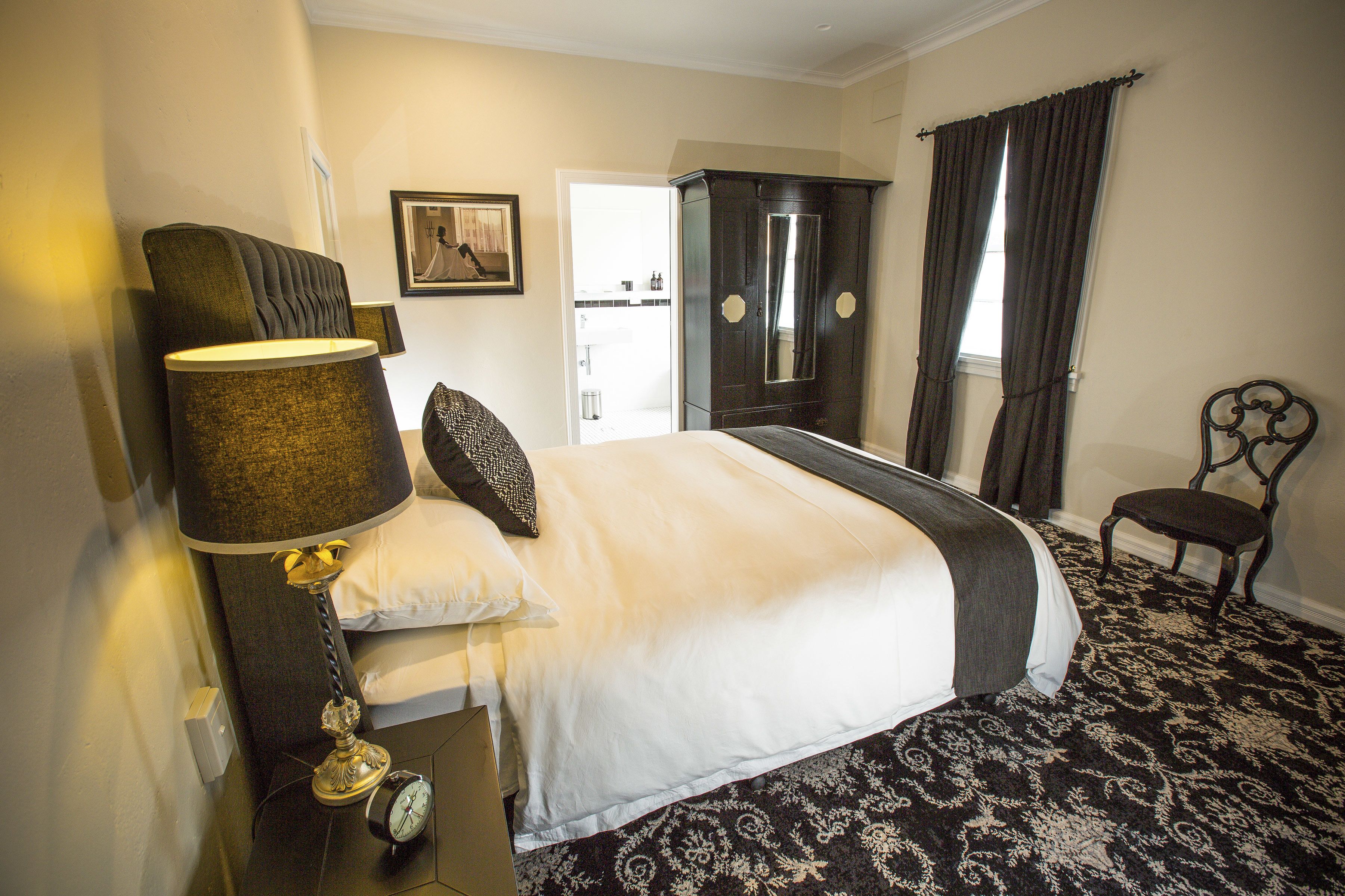 A hotel room with a large bed, a nightstand with a lamp, a chair, a wardrobe, and a framed picture on the wall.