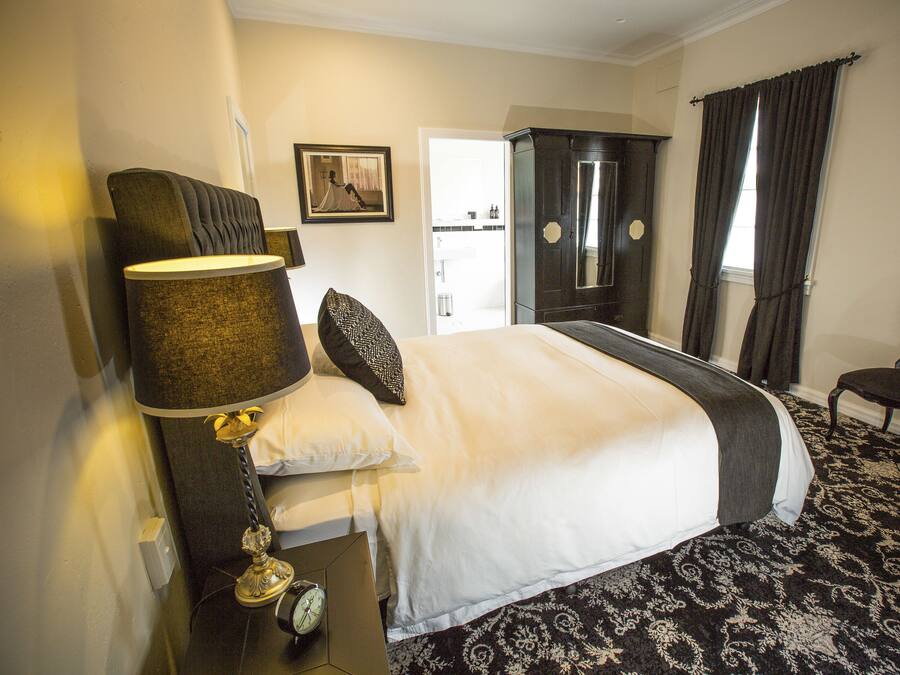 A hotel room with a large bed, a nightstand with a lamp, a chair, a wardrobe, and a framed picture on the wall.