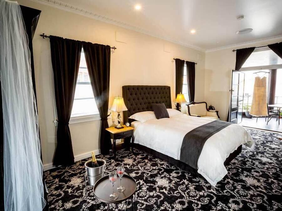 A hotel room with a large bed, two bedside lamps, a small table with a bottle and glasses, and a view of the outside through a door.