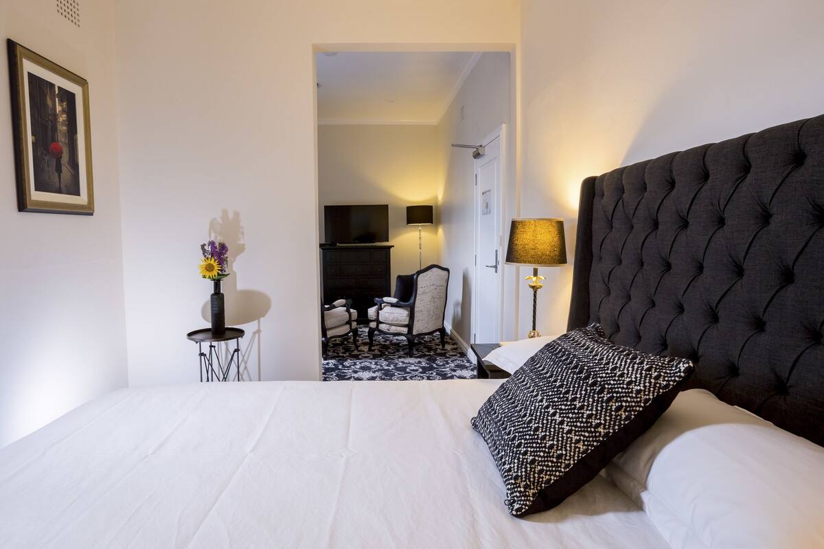 A hotel room with a large bed, a chair, a small table, and a TV in the background.