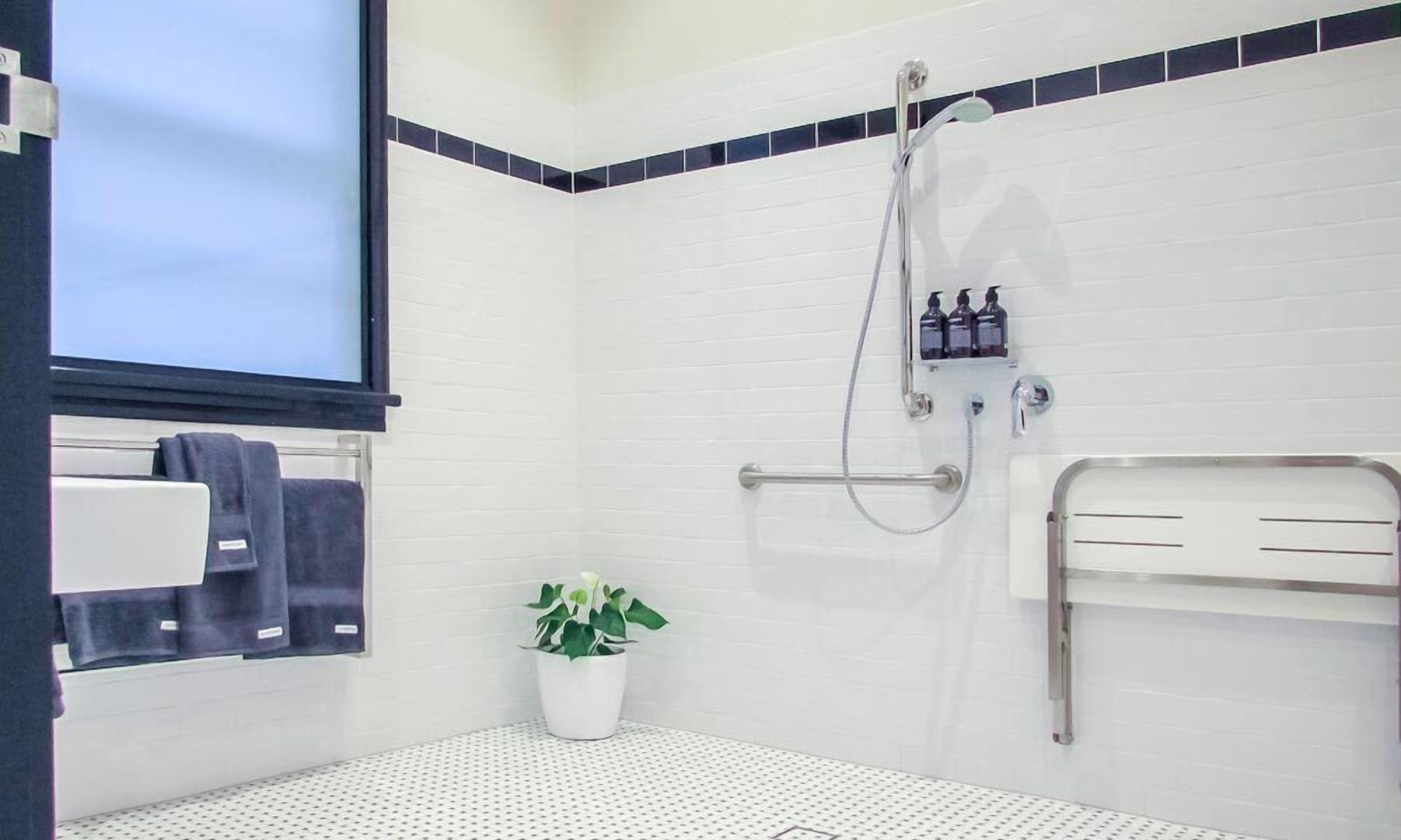 Un baño con paredes de azulejos blancos, ducha con manguera, toallero, planta en maceta e inodoro con barra de apoyo.