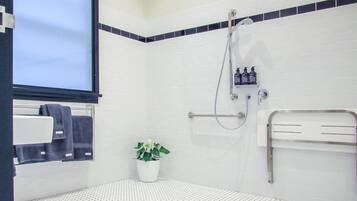 Un baño con paredes de azulejos blancos, ducha con manguera, toallero, planta en maceta e inodoro con barra de apoyo.
