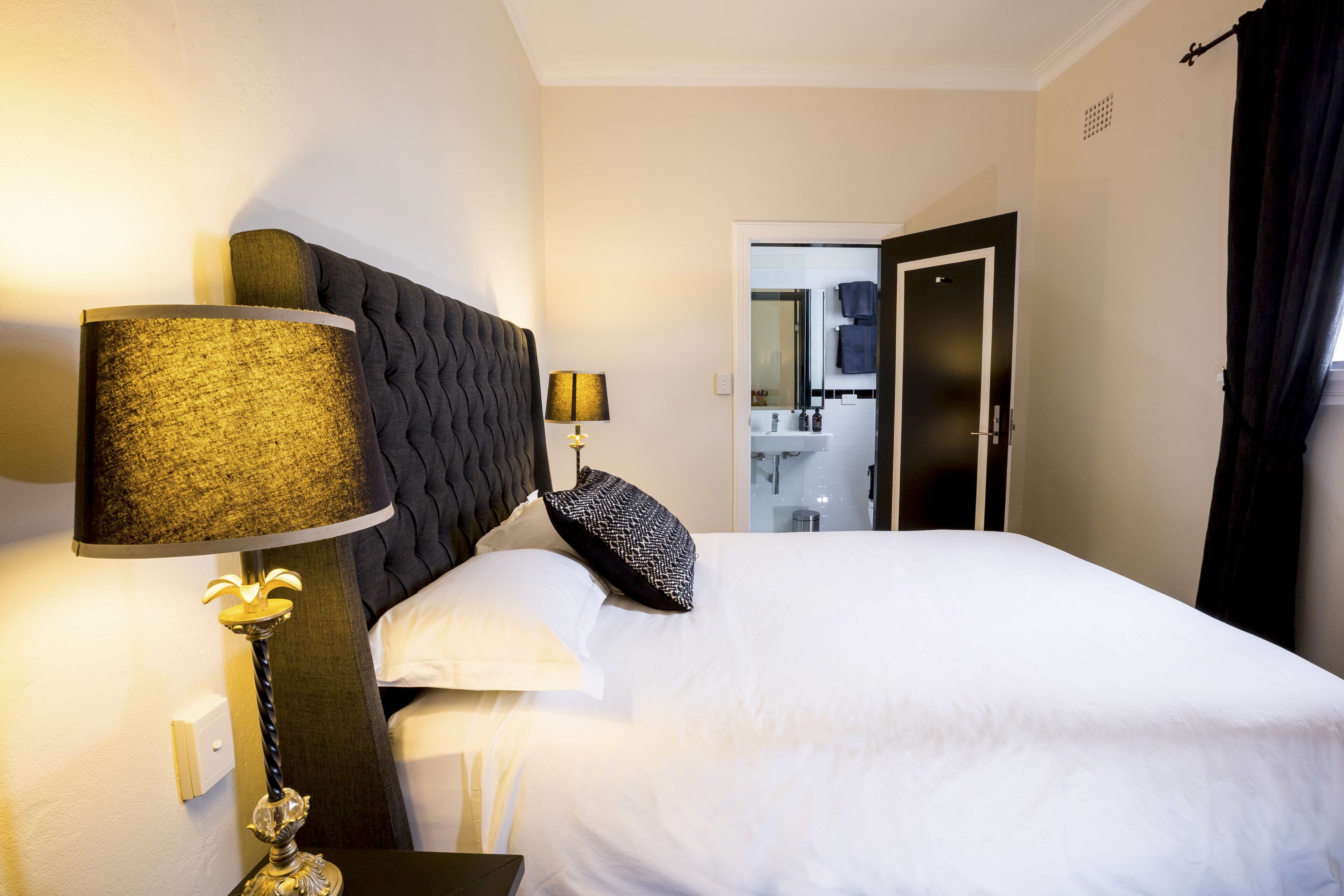 Une chambre d’hôtel avec un grand lit, une table de chevet avec une lampe, une tête de lit sombre et une vue sur la salle de bain par une porte ouverte.