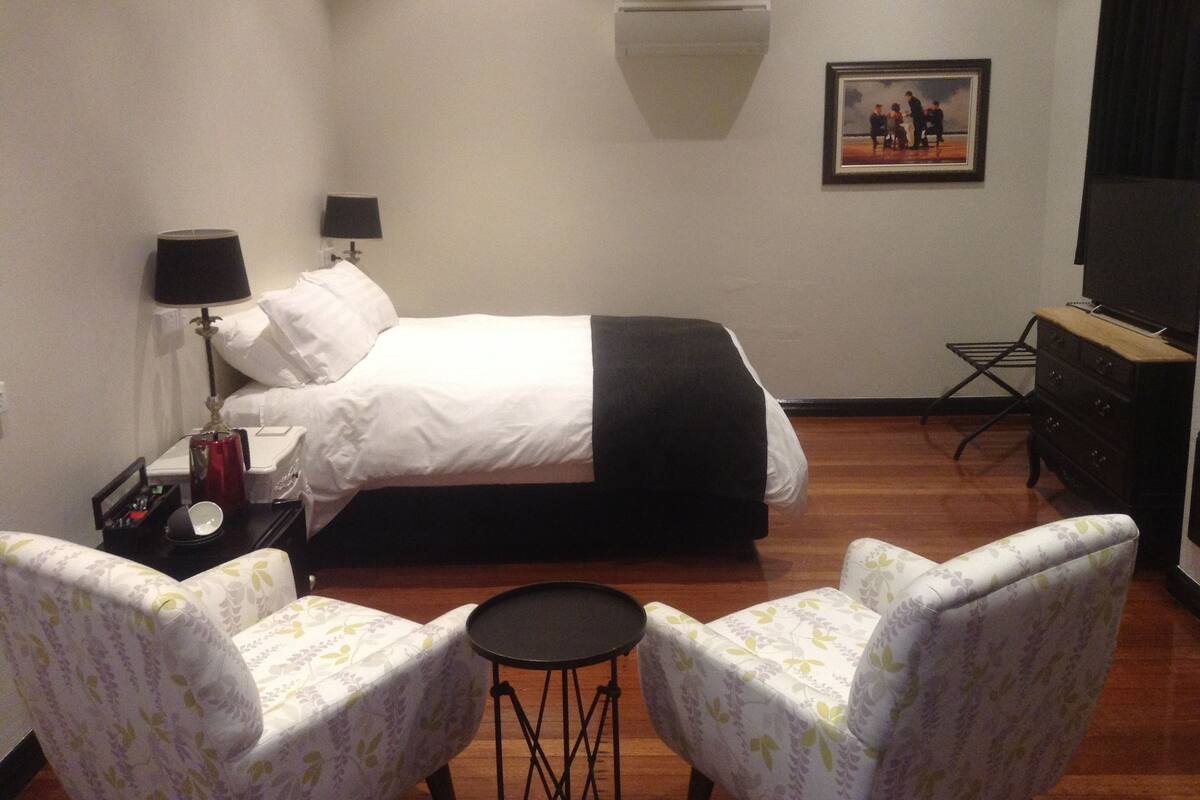 A hotel room with a bed, two armchairs, a small table, a TV, a painting on the wall, and a desk with a lamp.