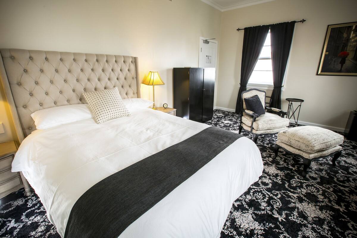 A hotel room with a large bed, a nightstand with a lamp, a wardrobe, a chair, and a small table.