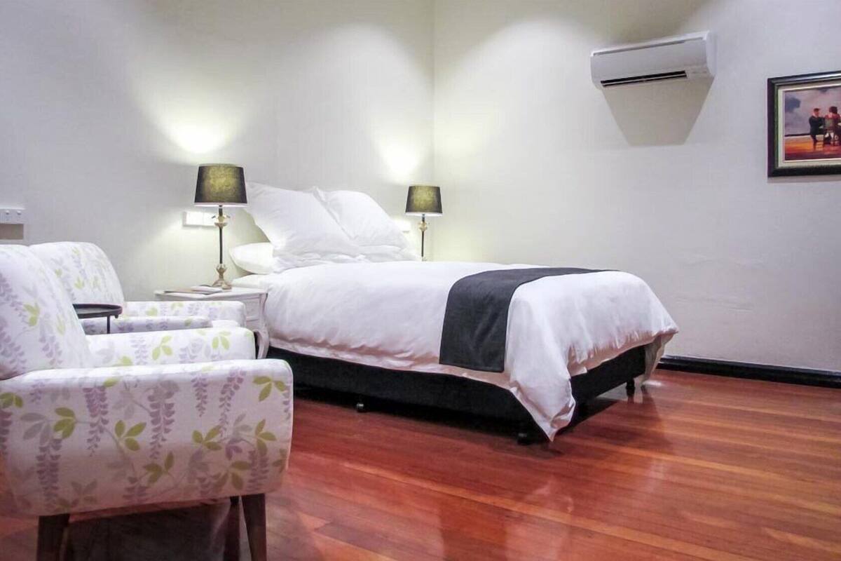 A hotel room with a bed, two bedside lamps, a chair, a small table, and an air conditioning unit.