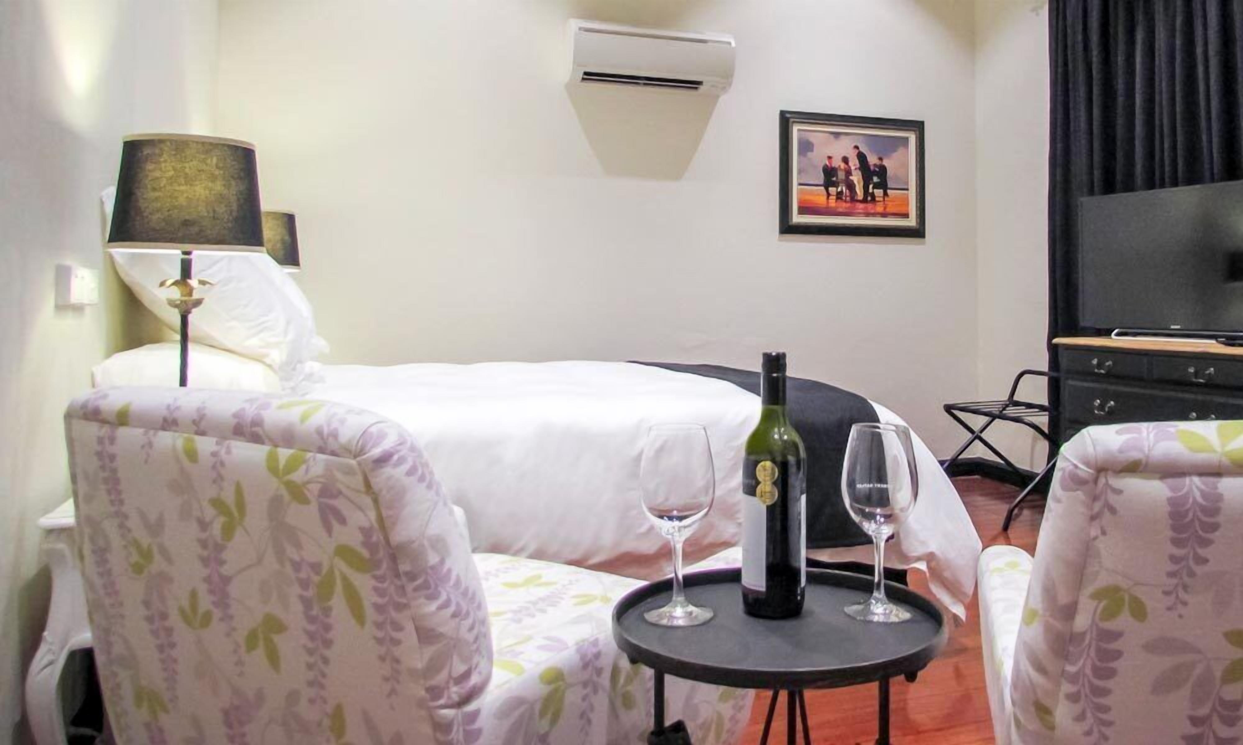 A hotel room with a bed, two chairs, a small table with a wine bottle and glass, a lamp, an air conditioner, and a framed picture on the wall.