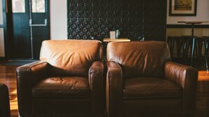 Un fauteuil en cuir marron se trouve dans une pièce dont le mur est recouvert d’un revêtement texturé foncé. Un tableau encadré est accroché au mur, et une porte est visible au fond.