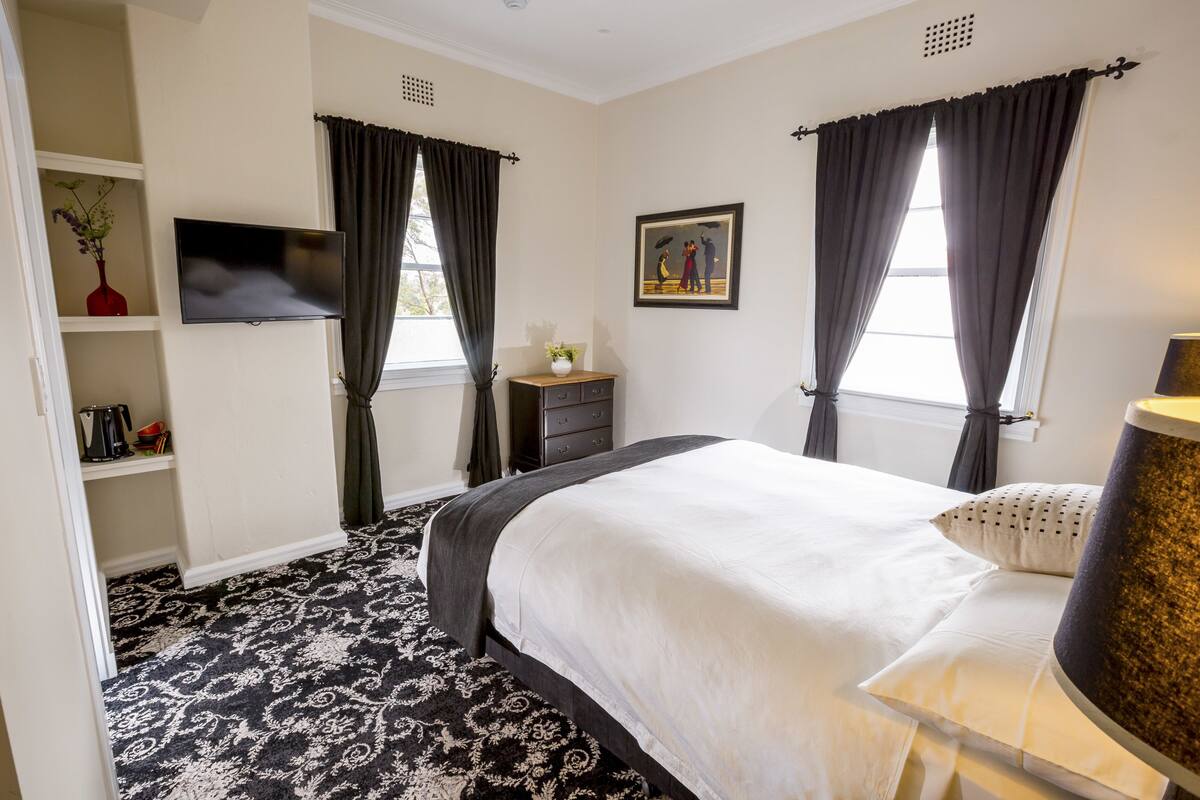 A bedroom with a large bed, a TV, a dresser, a nightstand, and a window with curtains.