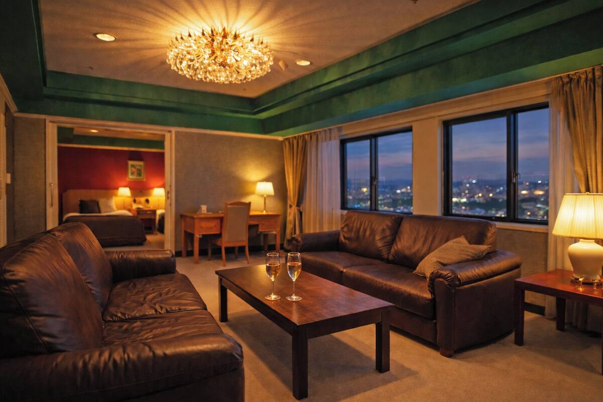A hotel room with a leather sofa set, a wooden coffee table, a chandelier, and a city view through large windows.