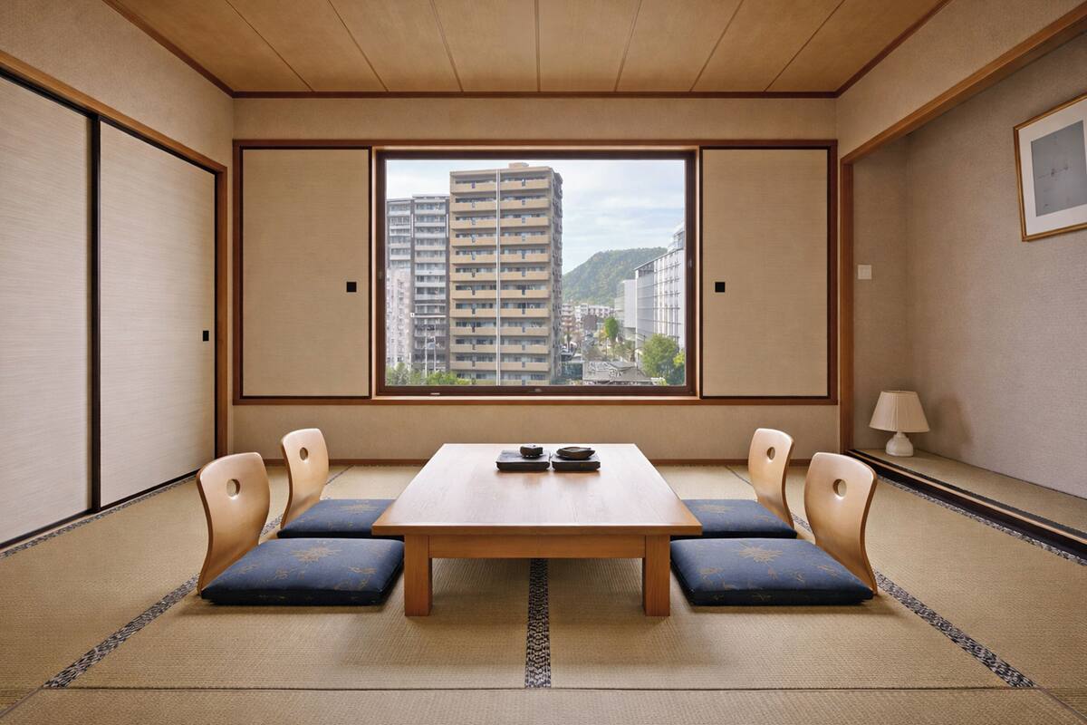 A traditional Japanese room with tatami flooring, a low wooden table, and a view of a cityscape through a large window.