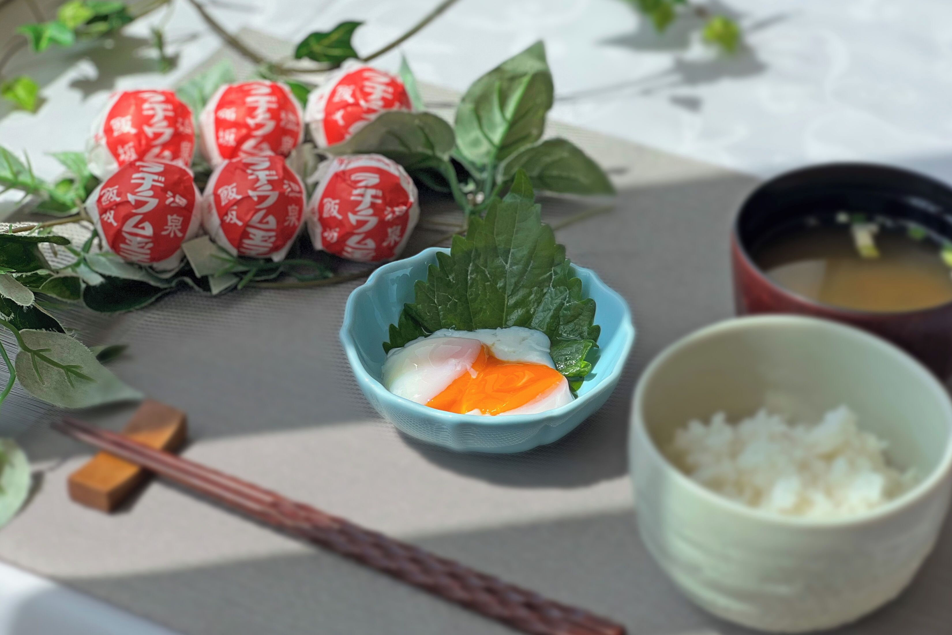 Una mesa con un tazón de arroz, una taza de té y objetos decorativos, incluyendo envoltorios rojos y blancos y hojas verdes.