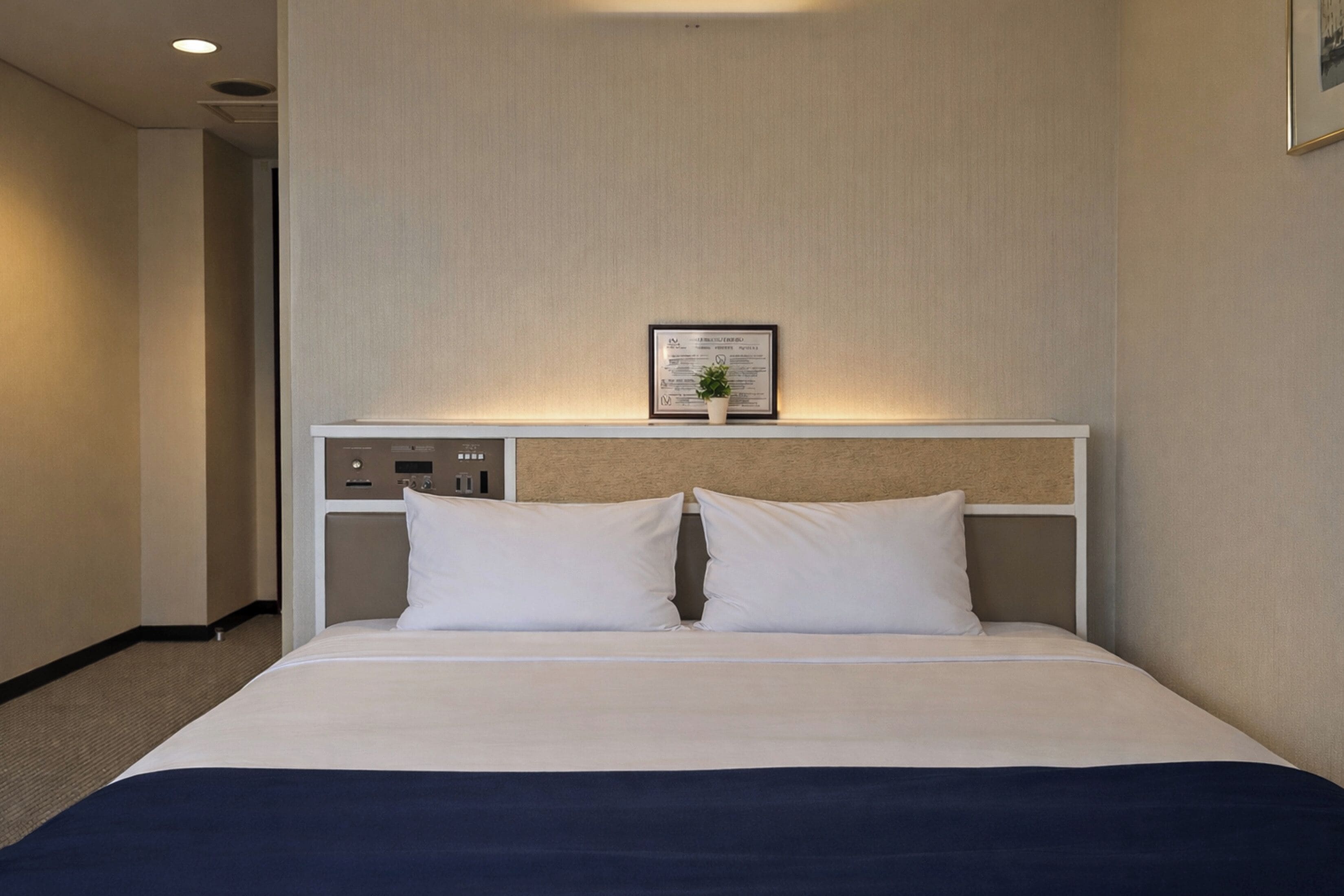 A hotel room with a bed, a headboard with controls, a framed picture, and a small plant.