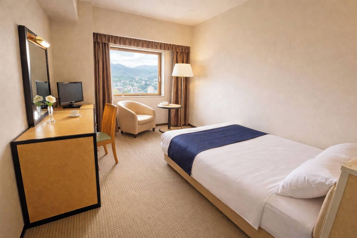 A hotel room with a bed, a desk with a computer, a chair, a lamp, and a window with a view.