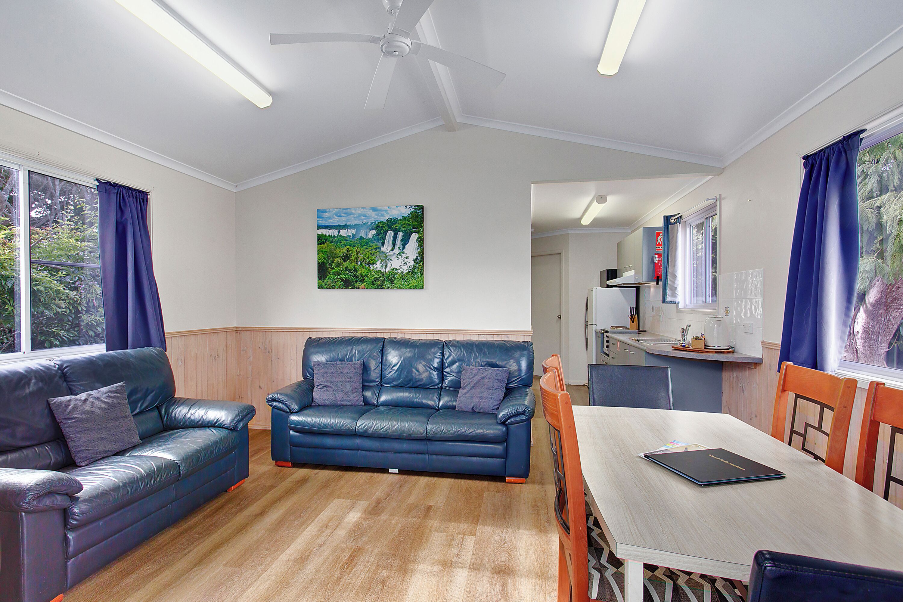 A living room with two blue leather sofas, a wooden dining table, and a ceiling fan.
