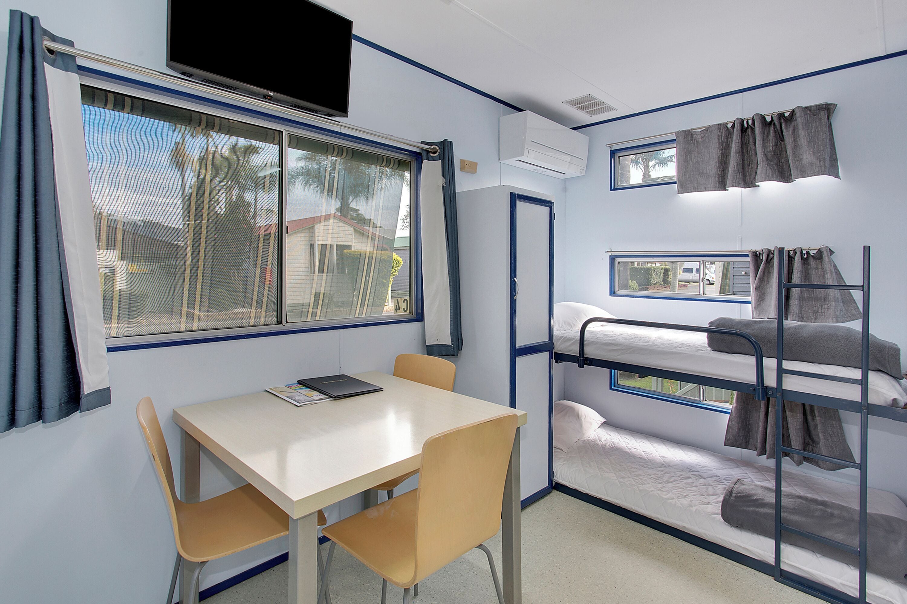 A bunk bed room with a table and chairs, a TV mounted on the wall, and a window with curtains.