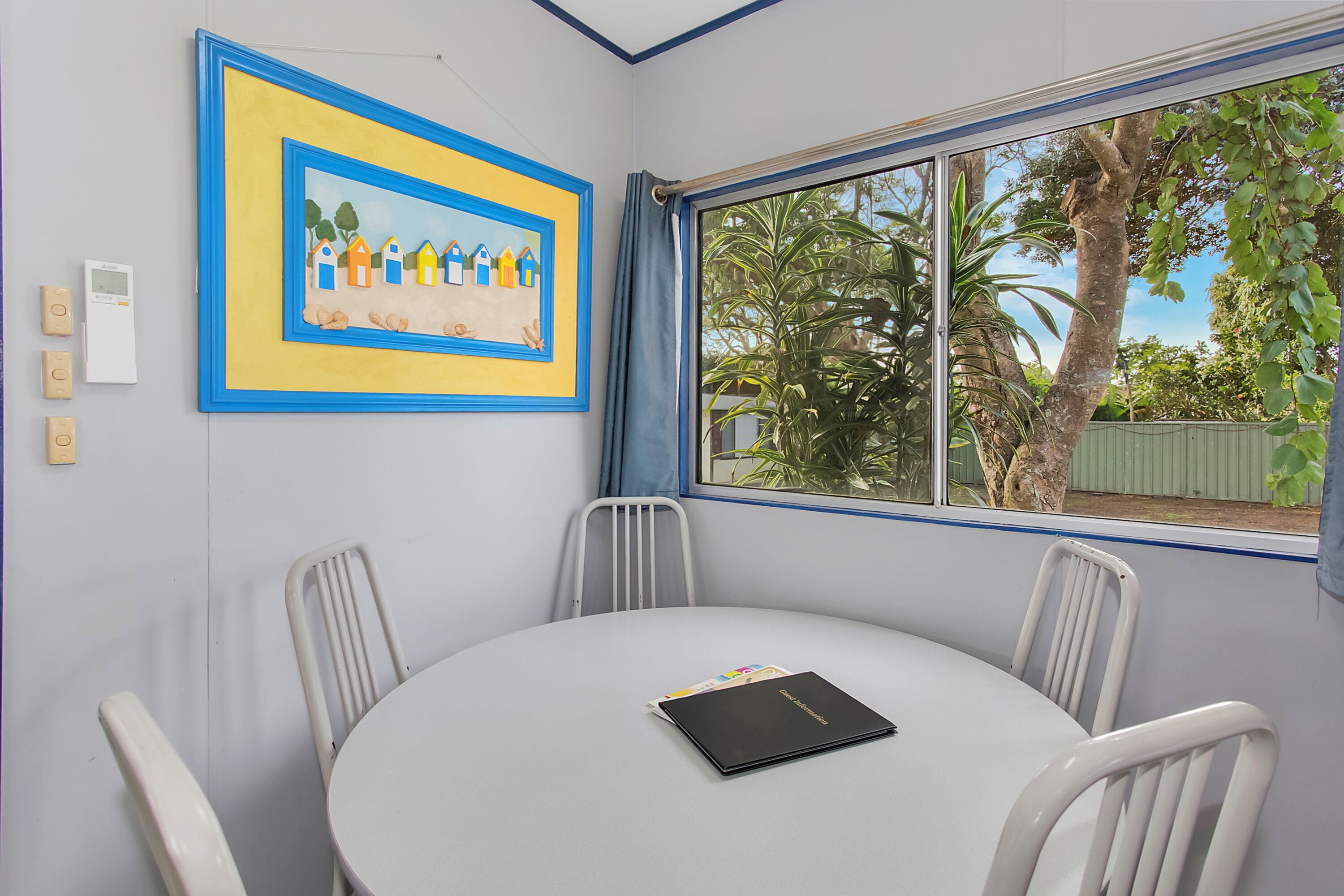 A conference room with a round table, four chairs, a framed painting, and a window with a view of trees and a fence.