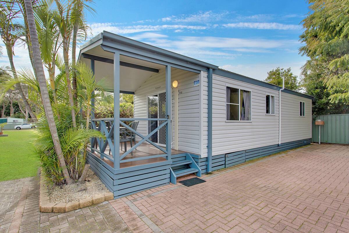 A single-story mobile home with a covered porch and a small deck, surrounded by a brick driveway and palm trees.