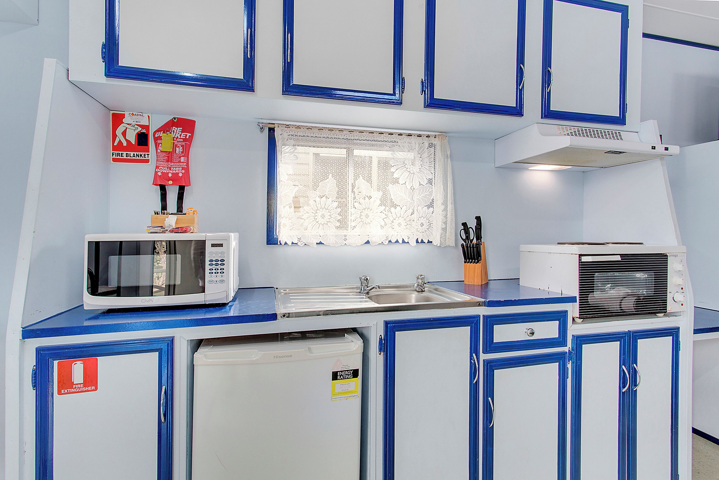 A kitchen with a microwave, toaster, and refrigerator. There is a fire extinguisher and a window with lace curtains.