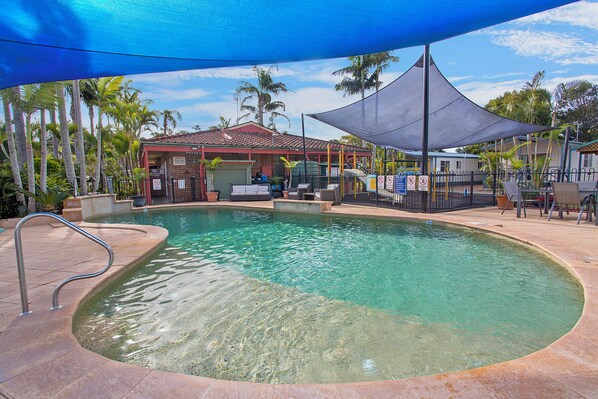 A clear swimming pool with a metal ladder, surrounded by palm trees and a shaded area with a roof. - North Haven Holiday Village (North Haven)