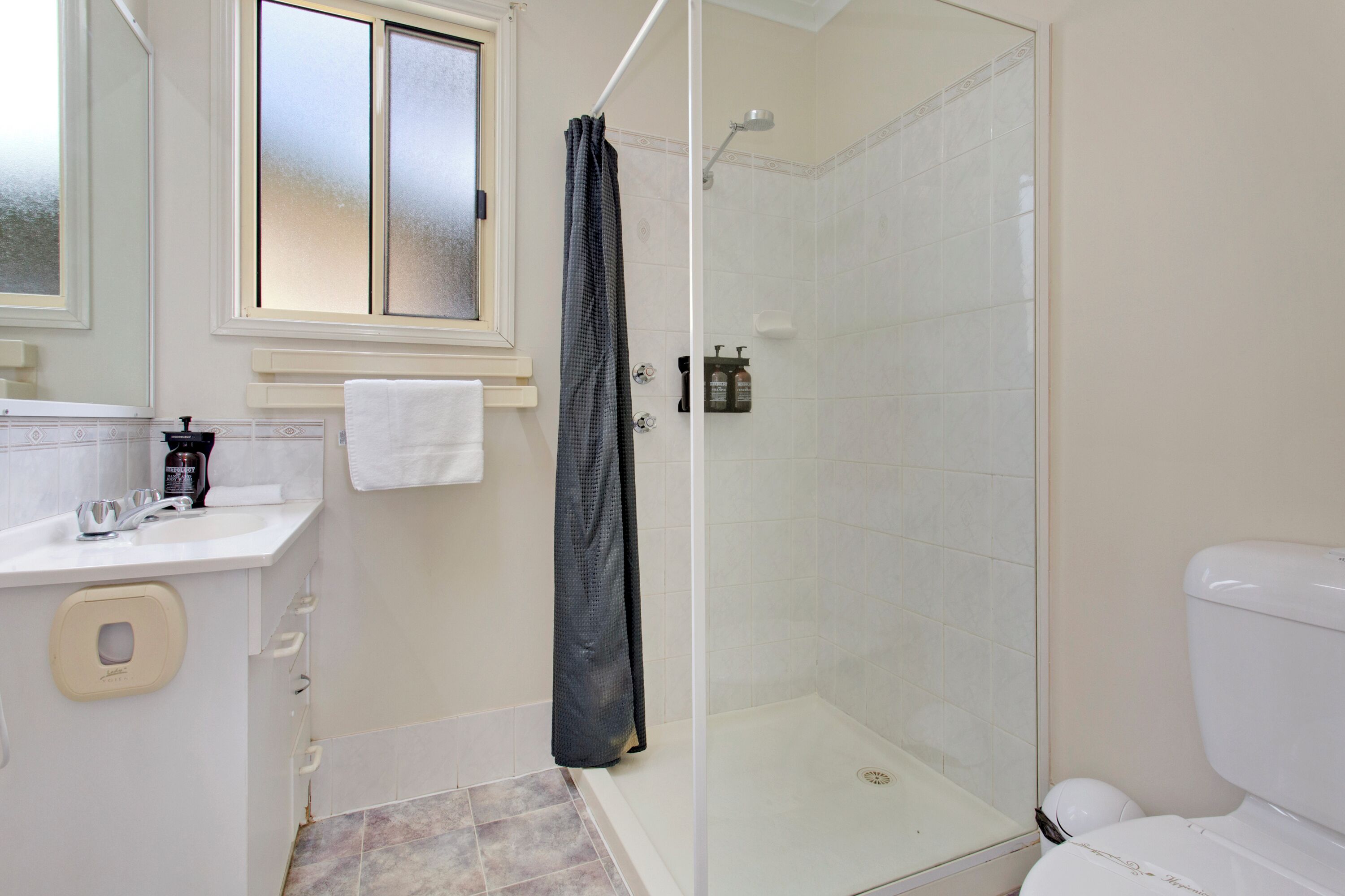A bathroom with a glass-enclosed shower, a white toilet, a sink with a soap dispenser, and a towel hanging on the wall.