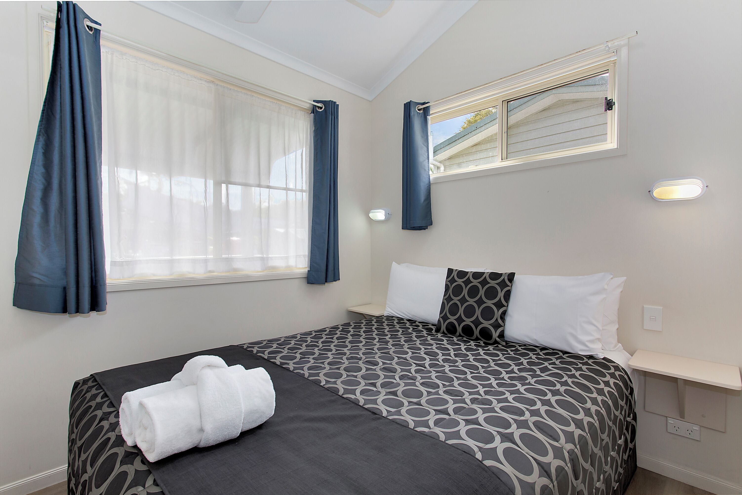 A neatly made bed with a patterned bedspread, a bedside table, and a window with curtains.