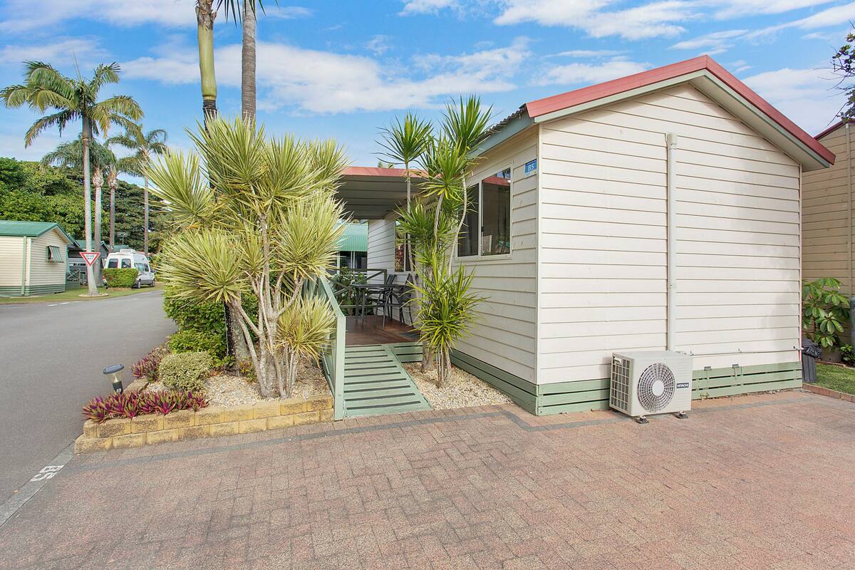 A small, single-story building with a red roof and beige siding, surrounded by palm trees and a paved driveway.