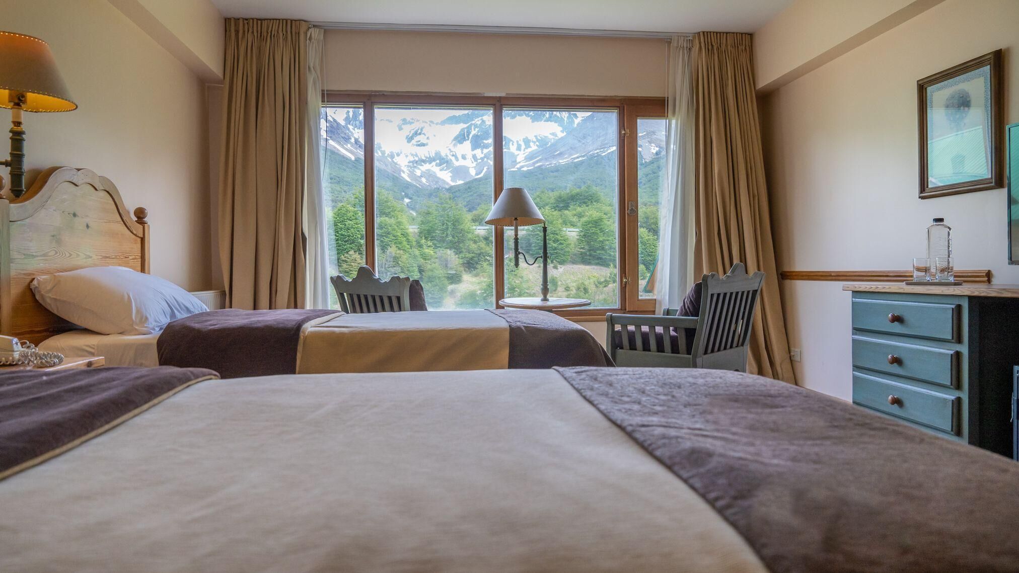 A hotel room with two beds, a desk, a chair, a lamp, and a view of mountains.