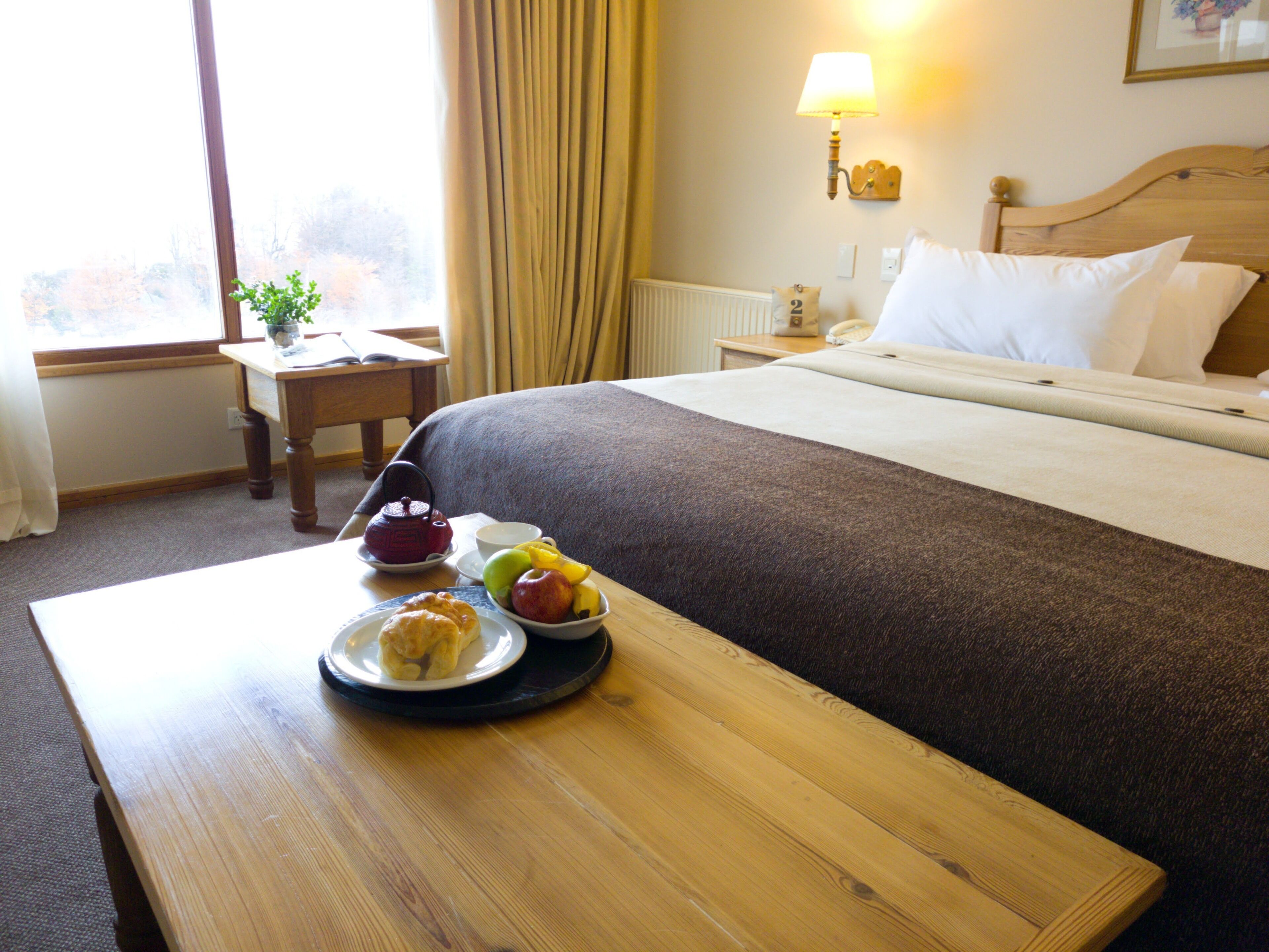 Habitación de hotel con una cama bien hecha, una mesita de noche con una bandeja de artículos de desayuno, una lámpara y una ventana con cortinas.