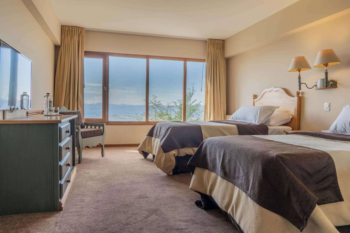 Ein Hotelzimmer mit zwei Betten, einem Schreibtisch, einem Schminktisch und einem großen Fenster mit Blick ins Freie.