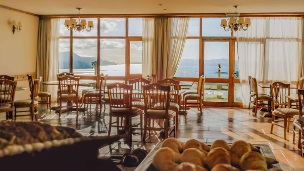 A dining area with wooden chairs and tables, a large window with a view of mountains and water, and a tray of bread.