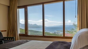 A hotel room with a large window offering a view of a coastal landscape, including mountains and a body of water.