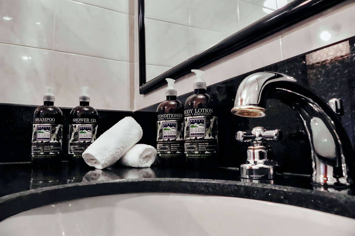 Ein Badezimmer mit schwarzem Waschtisch, einem Waschbecken und einem Spiegel. Auf dem Waschtisch stehen Flaschen mit Körperlotion und Shampoo.