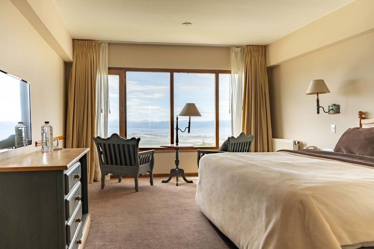 Ein Hotelzimmer mit Bett, Schreibtisch, zwei Stühlen, einer Lampe, Bergblick und einem fenster mit Vorhängen.