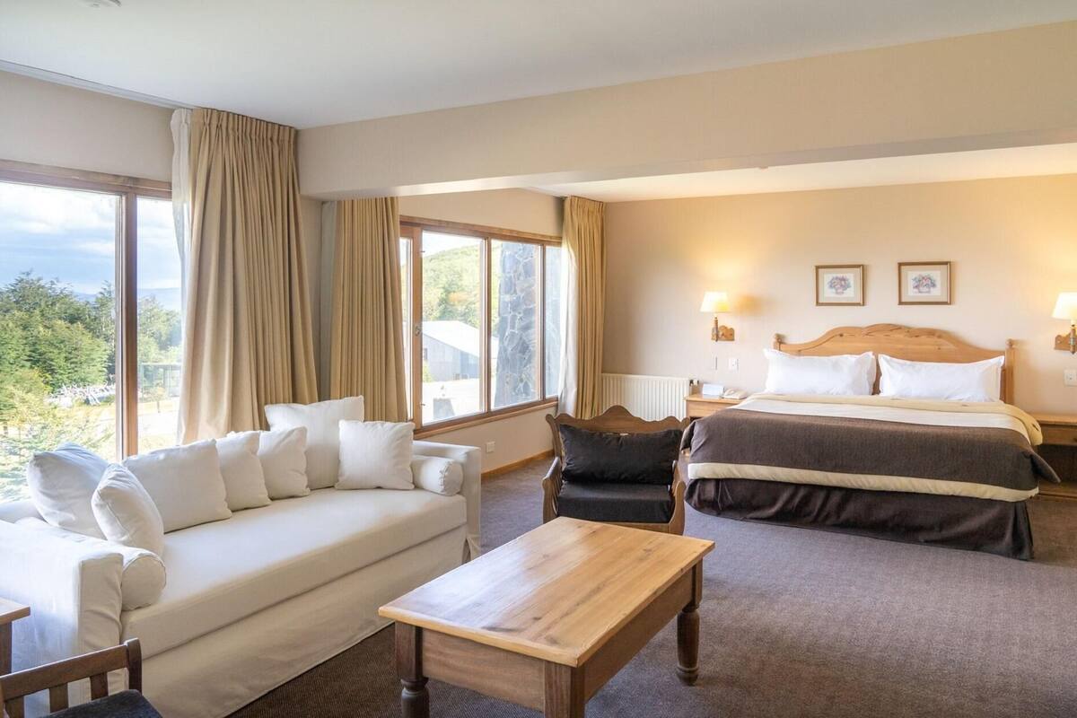 Ein Hotelzimmer mit einer Couch, einem Bett, einem Holztisch und großen Fenstern mit Vorhängen.