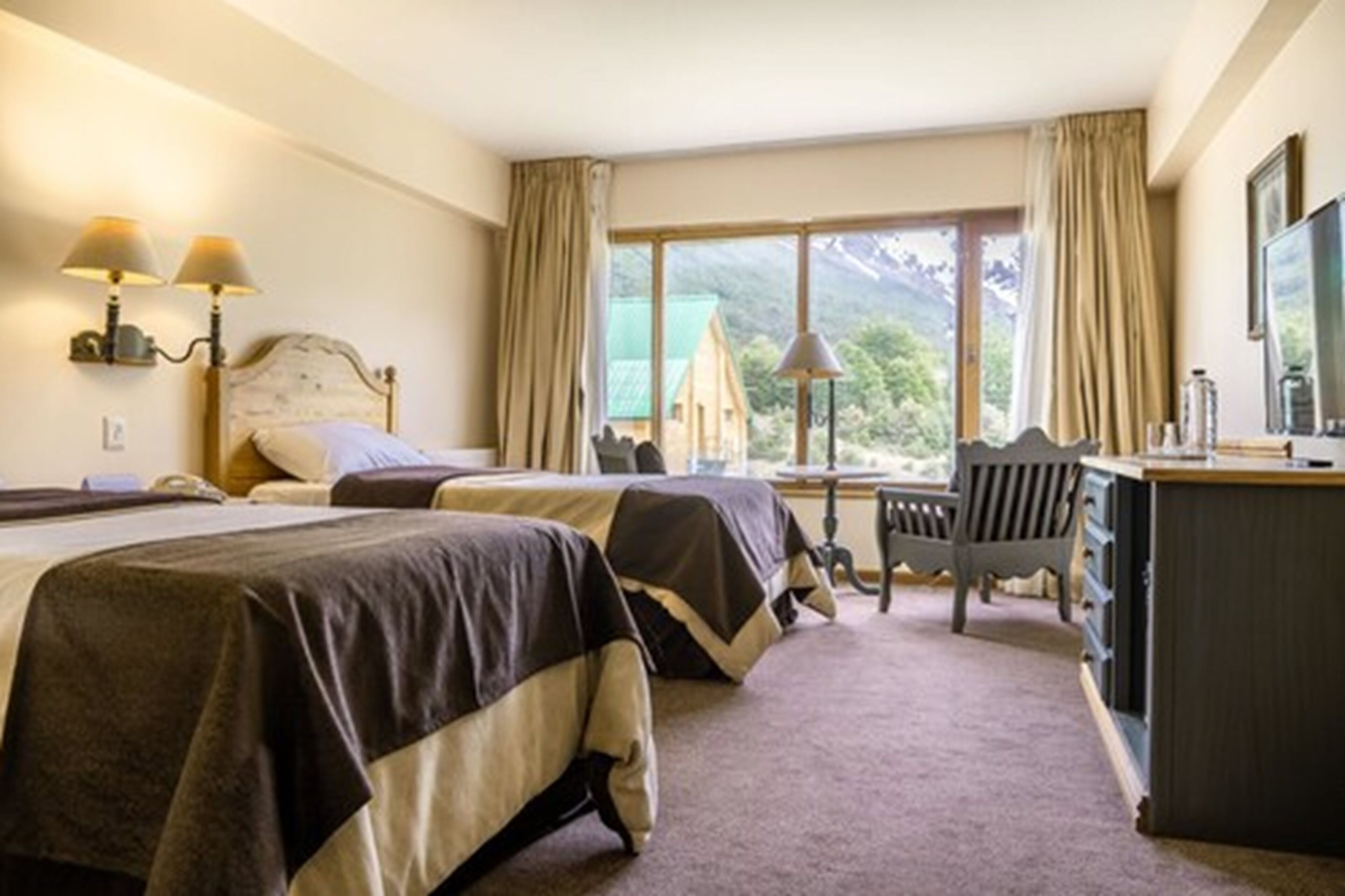 A hotel room with two beds, a desk, a chair, and a large window offering a mountain view.
